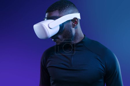 Foto de Atleta afroamericano sin emociones en ropa deportiva negra y auriculares blancos de realidad virtual de pie sobre fondo azul y mirando hacia abajo - Imagen libre de derechos