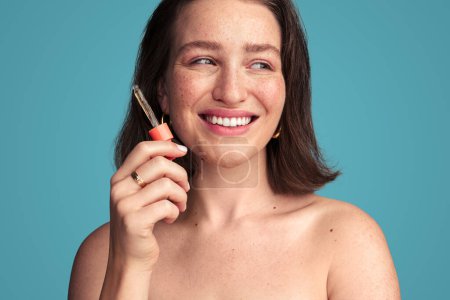 Foto de Modelo femenino joven sonriente con cabello oscuro y cuentagotas con suero para el tratamiento del cuidado de la piel contra el fondo turquesa - Imagen libre de derechos