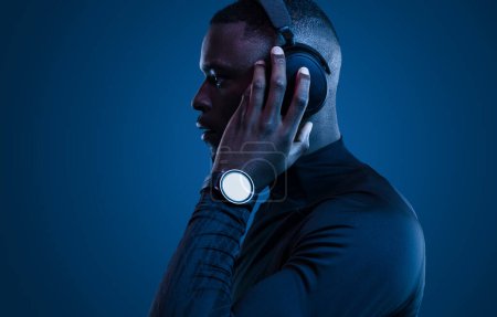 Photo for Side view of stylish young black male millennial with dark hair in activewear and smart watch, listening to song in modern wireless headphones and looking away against blue background - Royalty Free Image