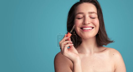 Foto de Mujer joven feliz con hombros desnudos y ojos cerrados sosteniendo la pipeta mientras aplica suero líquido en la cara contra el fondo azul - Imagen libre de derechos