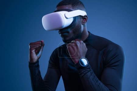 Téléchargez les photos : Jeune homme afro-américain barbu fort en vêtements de sport et montre intelligente debout en position de boxe tout en explorant le cyberespace dans les lunettes VR sur fond bleu - en image libre de droit