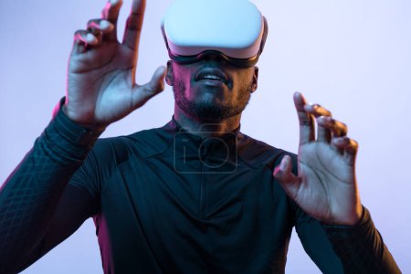 Téléchargez les photos : Concentré jeune homme afro-américain barbu en tenue futuriste noire et lunettes VR touchant écran invisible tout en explorant la réalité virtuelle sur fond violet - en image libre de droit