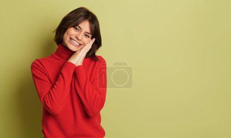 Foto de Joven hembra tímida positiva en cuello alto rojo con las manos apretadas tocando la mejilla mientras sonríe y mira a la cámara contra un fondo vívido - Imagen libre de derechos