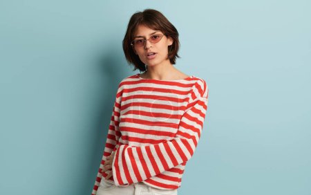 Photo for Confident young female in trendy striped outfit and sunglasses looking at camera while standing against blue background - Royalty Free Image