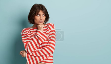 Foto de Atractiva joven hembra en traje de moda pensando y dudando mientras mira hacia otro lado en el espacio de copia sobre fondo azul - Imagen libre de derechos
