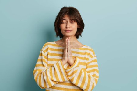 Photo for Calm young female with short hair in yellow striped sweater standing near blue wall and meditating with closed eyes - Royalty Free Image