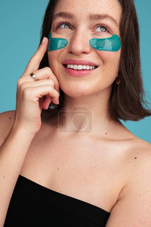 Photo for Crop young attractive female model touching patch under eye while having beauty procedure against turquoise background looking up - Royalty Free Image