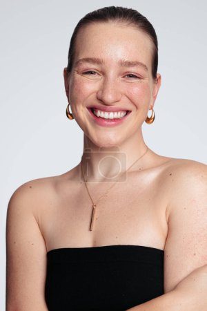 Téléchargez les photos : Jeune femme positive aux épaules nues souriant et regardant la caméra sur fond blanc représentant le concept de beauté et le maquillage naturel - en image libre de droit