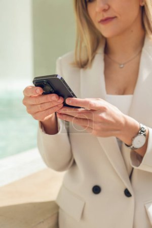 Téléchargez les photos : Crop femme employée en veste blanche à la mode navigation téléphone mobile près de la fenêtre dans la salle de lumière - en image libre de droit