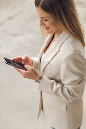 Photo for From above side view of crop cheerful adult businesswoman with long hair in stylish suit, smiling while messaging on smartphone standing on street in daytime - Royalty Free Image