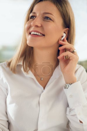 Téléchargez les photos : Jeune femme d'affaires positive en chemise blanche touchant les écouteurs tout en écoutant de la musique sur fond flou pendant la pause du travail - en image libre de droit