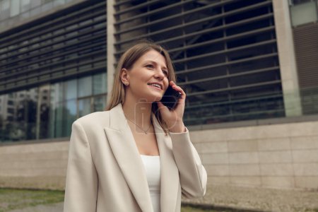Foto de Mujer joven feliz en traje elegante sonriendo y teniendo conversación telefónica mientras está de pie en la calle de la ciudad cerca del edificio moderno - Imagen libre de derechos