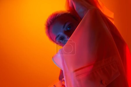 Photo for Side view of confident young female model with short blond hair in transparent raincoat looking at camera against orange background in neon lights - Royalty Free Image