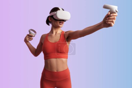 Téléchargez les photos : Femme sportive dans un casque VR et avec des contrôleurs ouvrant la bouche tout en interagissant avec la réalité virtuelle sur fond de dégradé - en image libre de droit