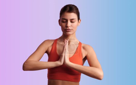 Téléchargez les photos : S'adapter jeune modèle féminin hispanique calme avec les cheveux foncés en vêtements de sport méditant avec les yeux fermés et les mains namaste, tout en pratiquant le yoga sur fond deux couleurs - en image libre de droit