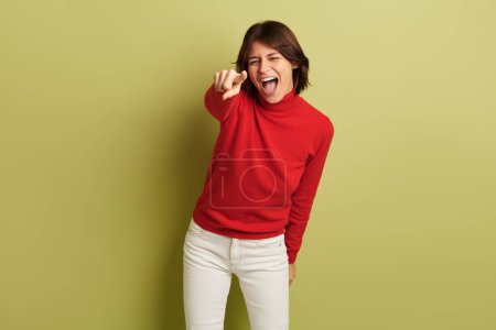 Photo for Delighted young Hispanic female model with dark hair in red turtleneck screaming expressively and pointing at camera against green background in studio - Royalty Free Image