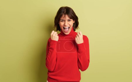 Foto de Mujer joven feliz en jersey de cuello alto rojo celebrando una victoria, mientras sonríe sobre fondo verde - Imagen libre de derechos