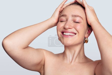 Téléchargez les photos : Joyeux jeune femme avec un maquillage naturel propre fixant les cheveux avec les yeux fermés sur fond blanc en studio - en image libre de droit