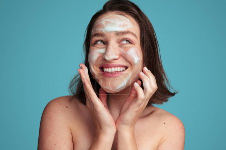 Photo for Cheerful young female model with dark hair and bare shoulders smiling happily and looking away wile washing face with foam cleanser against blue background - Royalty Free Image