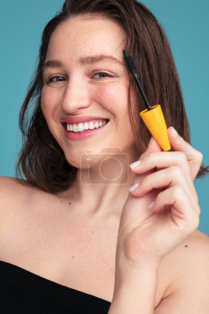 Foto de Confiado modelo femenino joven con el pelo oscuro y pecas sonriendo y mirando a la cámara mientras se aplica rímel sobre fondo azul - Imagen libre de derechos