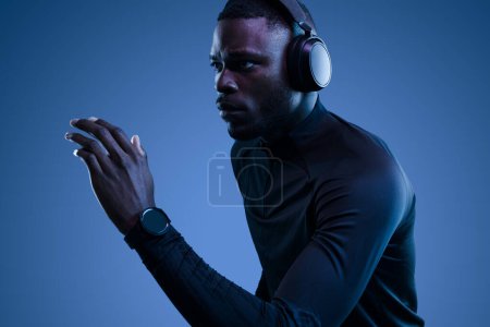 Foto de Esprinter masculino afroamericano concentrado en ropa deportiva negra y reloj inteligente escuchando música en auriculares inalámbricos mientras se ejecuta sobre fondo azul - Imagen libre de derechos