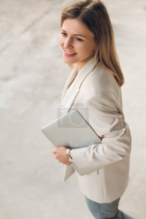 Téléchargez les photos : Angle élevé de jeune femme d'affaires positive en tenue décontractée intelligente marchant dans la rue de la ville avec netbook - en image libre de droit