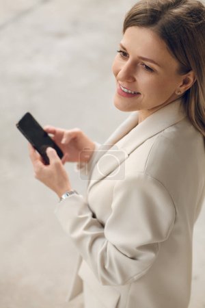 Foto de Desde arriba de la mujer adulta sonriente en chaqueta de pie en la calle y el uso de un teléfono inteligente mientras mira hacia otro lado - Imagen libre de derechos