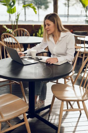 Téléchargez les photos : Vue latérale d'une jeune pigiste en chemise blanche assise à une table ronde dans un café et travaillant en ligne sur un projet à l'aide d'un ordinateur portable - en image libre de droit