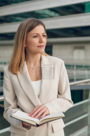 Téléchargez les photos : Jeune femme d'affaires en tenue décontractée intelligente debout à l'extérieur du centre d'affaires avec planificateur et regardant loin réfléchie - en image libre de droit