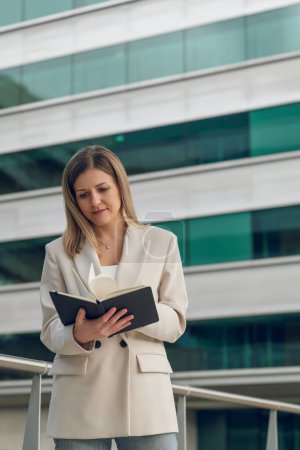 Téléchargez les photos : Jeune entrepreneure confiante dans une tenue élégante debout près d'un immeuble de bureaux moderne tout en lisant un journal intime en ville pendant le travail - en image libre de droit