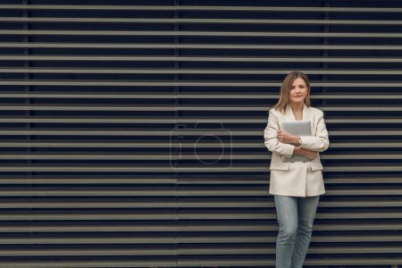 Téléchargez les photos : Femme d'affaires confiante dans une tenue élégante tenant un ordinateur portable regardant la caméra tout en se tenant debout contre un fond mural rayé pendant la journée - en image libre de droit