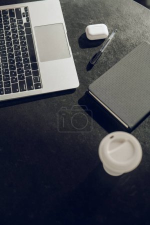 Foto de Alto ángulo de netbook moderno colocado en el escritorio negro con auriculares de la pluma caso copybook y taza de papel - Imagen libre de derechos