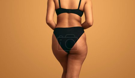 Back view of crop anonymous plump female in black panties and bra with imperfect skin standing against orange background