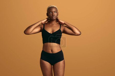 Téléchargez les photos : Jeune modèle féminin afro-américain réfléchi avec cheveux courts teints en sous-vêtements noirs touchant le cou et regardant loin sur fond beige - en image libre de droit