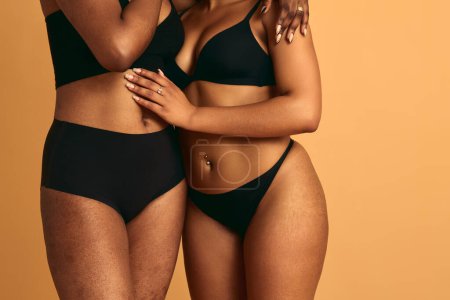 Photo for Crop anonymous ethnic plus size lesbian couple in black underwear embracing each other against beige background - Royalty Free Image