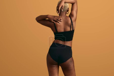 Foto de Vista posterior de la mujer africana en lencería negra con estrías en el cuerpo tocando el hombro mientras está de pie sobre fondo naranja - Imagen libre de derechos