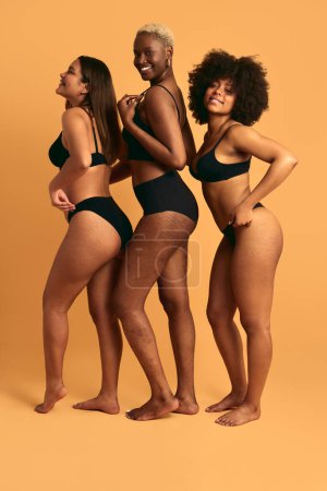 Foto de Vista lateral de cuerpo completo de alegres modelos femeninos jóvenes multiétnicos en ropa interior negra de pie sobre fondo amarillo y sonriendo felizmente en el estudio - Imagen libre de derechos
