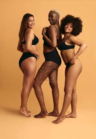 Photo for Full body of young multiracial girlfriends in black lingerie standing against orange background smiling and looking at camera - Royalty Free Image