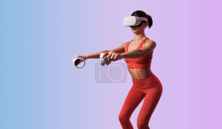 Photo for Slim female wearing VR headset and red sportswear holding controllers while doing squats against gradient background - Royalty Free Image