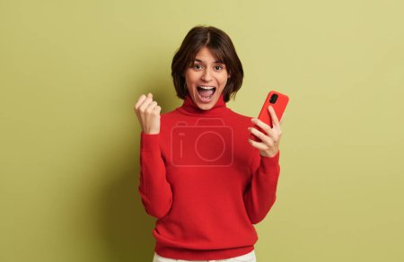 Foto de Joven mujer alegre con el pelo corto en cuello alto rojo de pie cerca de fondo verde con teléfono inteligente y celebrando el logro - Imagen libre de derechos