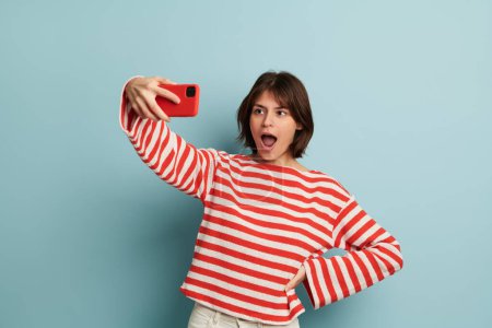Photo for Confident young female blogger with dark hair in red and white striped blouse taking selfie with opened mouth on smartphone , while standing against blue background with hand on waist - Royalty Free Image