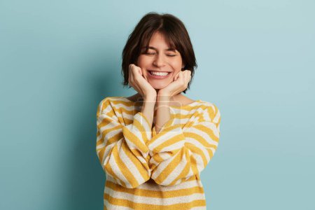 Optimistic young Hispanic female model with dark hair in striped white and yellow blouse, smiling brightly with closed eyes and hands on cheeks against blue background
