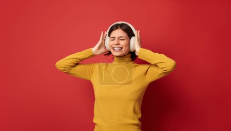 Foto de Mujer satisfecha en cuello alto amarillo cerrando los ojos y tocando auriculares inalámbricos en la cabeza mientras escucha música sobre fondo rojo - Imagen libre de derechos