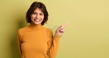 Foto de Modelo femenino étnico joven positivo con cabello oscuro en cuello alto amarillo sonriendo brillantemente y mirando a la cámara mientras apunta hacia el estudio verde - Imagen libre de derechos