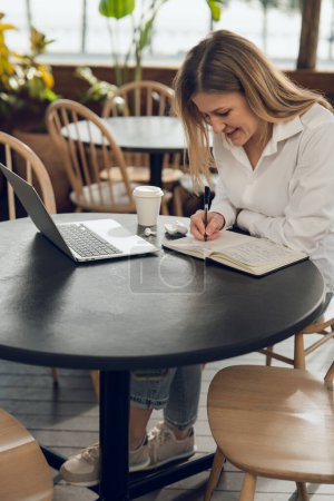 Téléchargez les photos : Jeune femme pigiste en vêtements décontractés assis à table avec ordinateur portable et écrit dans un journal tout en travaillant sur un projet à distance dans un café - en image libre de droit