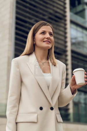 Photo for Low angle of positive young businesswoman in beige jacket standing outside modern business center with takeaway hot drink and looking away - Royalty Free Image