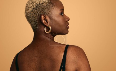 Photo for Back view of pensive young African American female model with short curly hair and earrings, wearing black top looking over shoulder against beige background in studio - Royalty Free Image