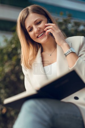Téléchargez les photos : Angle bas de jeune femme d'affaires souriante en vêtements décontractés intelligents assis à l'extérieur et parlant via un téléphone mobile tout en lisant des informations dans le planificateur - en image libre de droit
