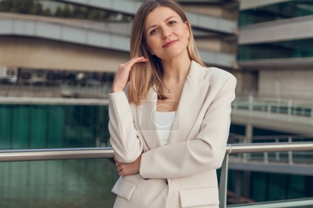 Téléchargez les photos : Femme d'affaires positive confiante en costume formel debout sur le balcon dans le quartier des affaires et toucher les cheveux tout en regardant la caméra - en image libre de droit