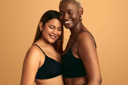 Téléchargez les photos : Sensuelles jeunes copines multiraciales en lingerie noire avec les yeux fermés debout près de l'autre sur fond orange - en image libre de droit